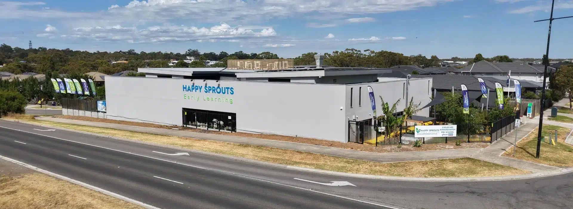 Exterior view of the Happy Sprouts Early Learning preschool building along a curved street, with blue and green signage and a fenced playground area outside.