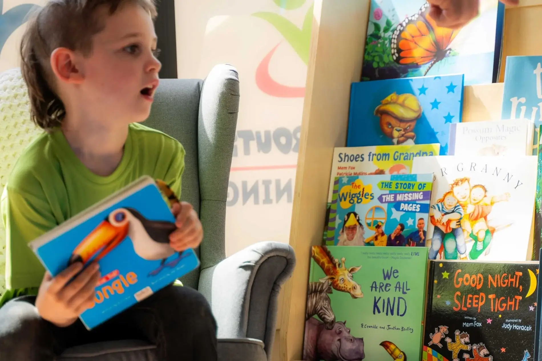 Side view of a boy in a green shirt sitting on a sofa, reading a blue children's book as colorful picture books line a shelf nearby.