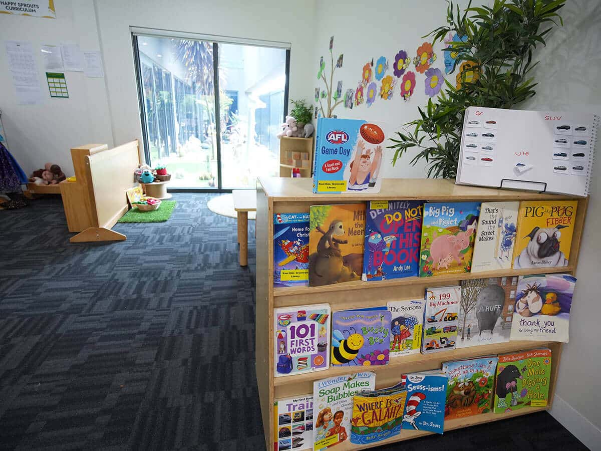 Children's library corner with a wooden bookshelf of colorful picture books, wall flower decorations, and a sliding glass door to the yard.