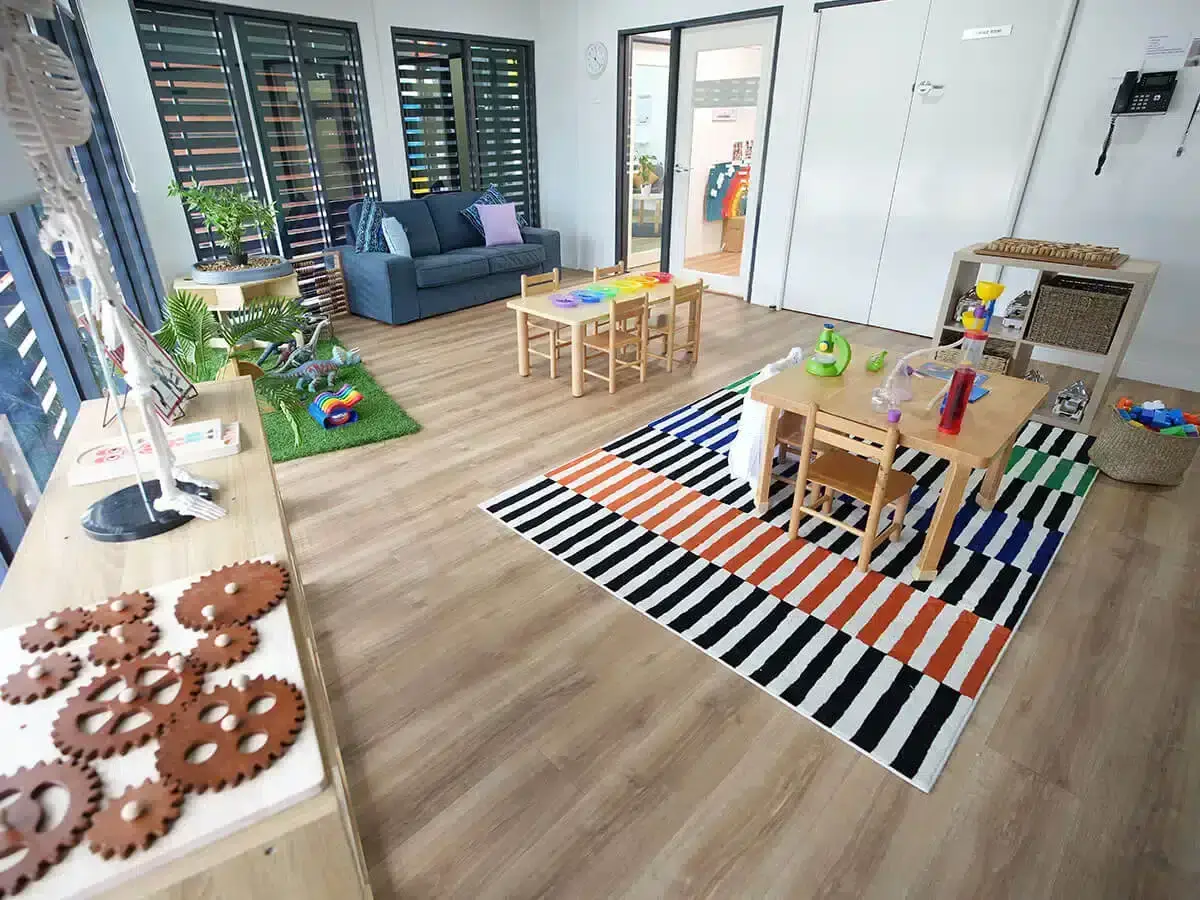 Bright children's playroom with small wooden tables and chairs, colorful toys, and a striped rug on wood flooring.