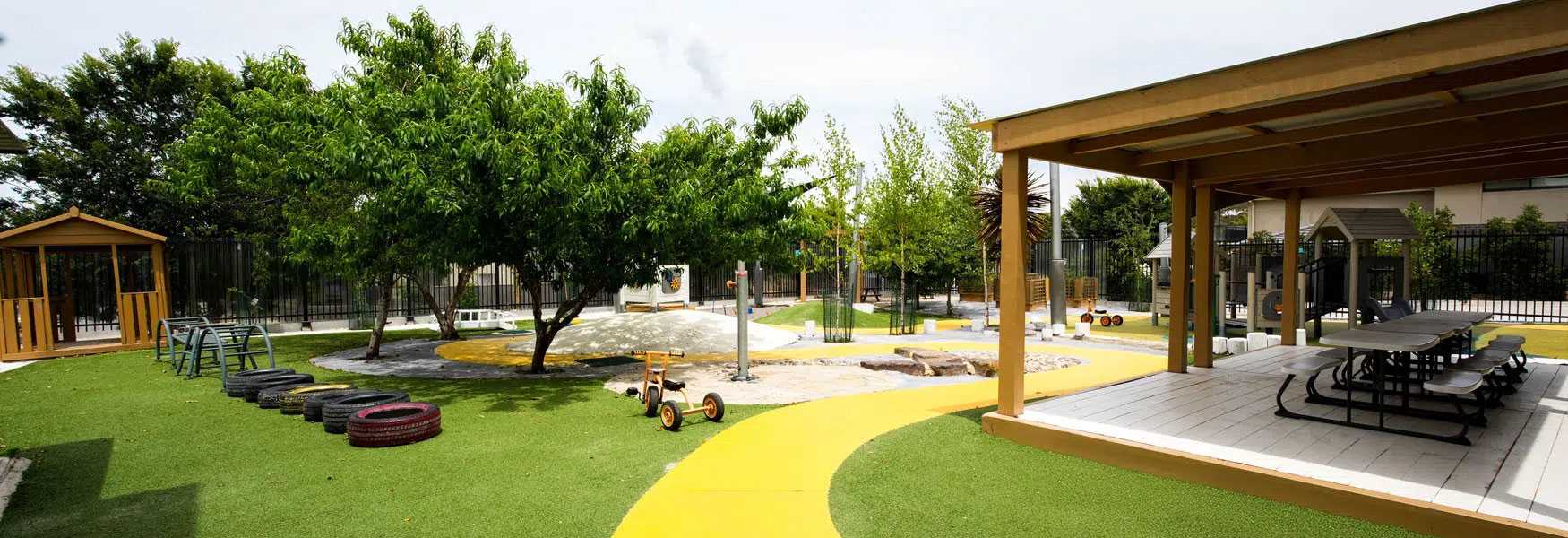 Playground with green turf, trees, a wooden playhouse, tire obstacles, and a shaded pavilion with picnic tables.