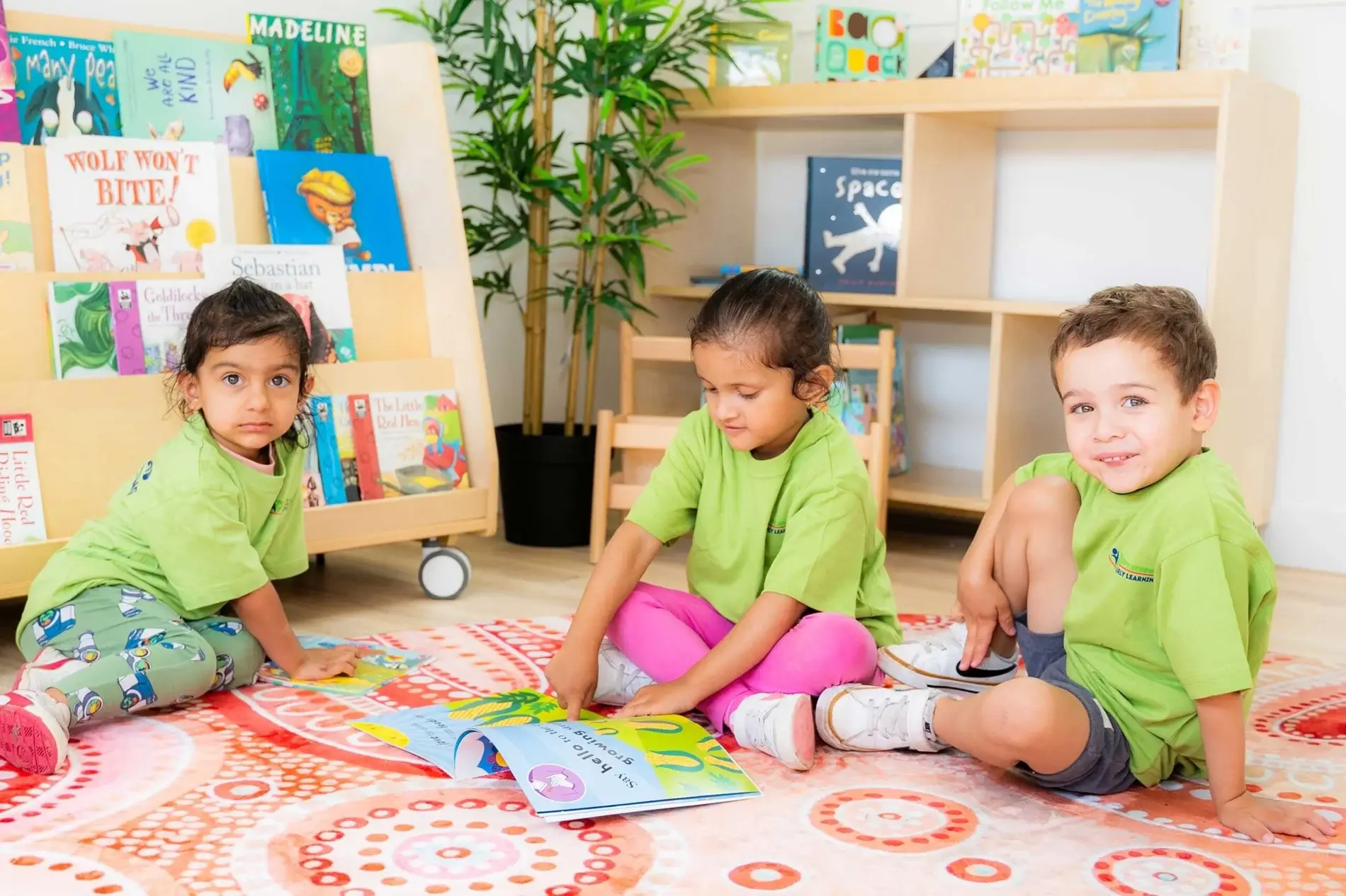 Three children in green shirts sit on a colorful rug, reading together in a bright library with child-friendly bookshelves behind them.