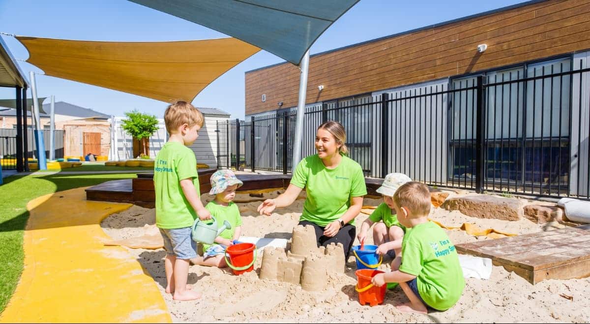 Caregiver and four kids in green shirts playing in a sandbox under shade sails in a sunny outdoor area.
